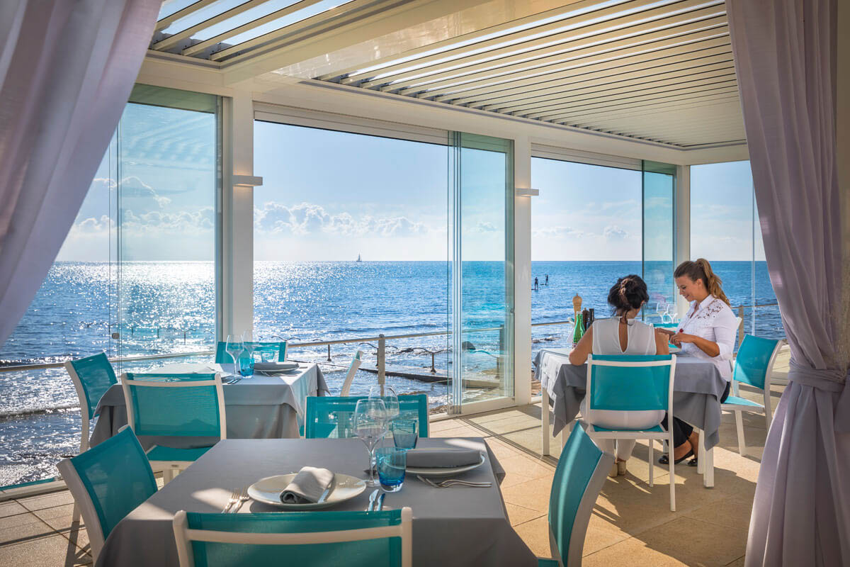 Zwei Personen sitzen an einem Tisch in einem Restaurant mit großen Fenstern und Blick auf das Meer bei Sonnenschein.