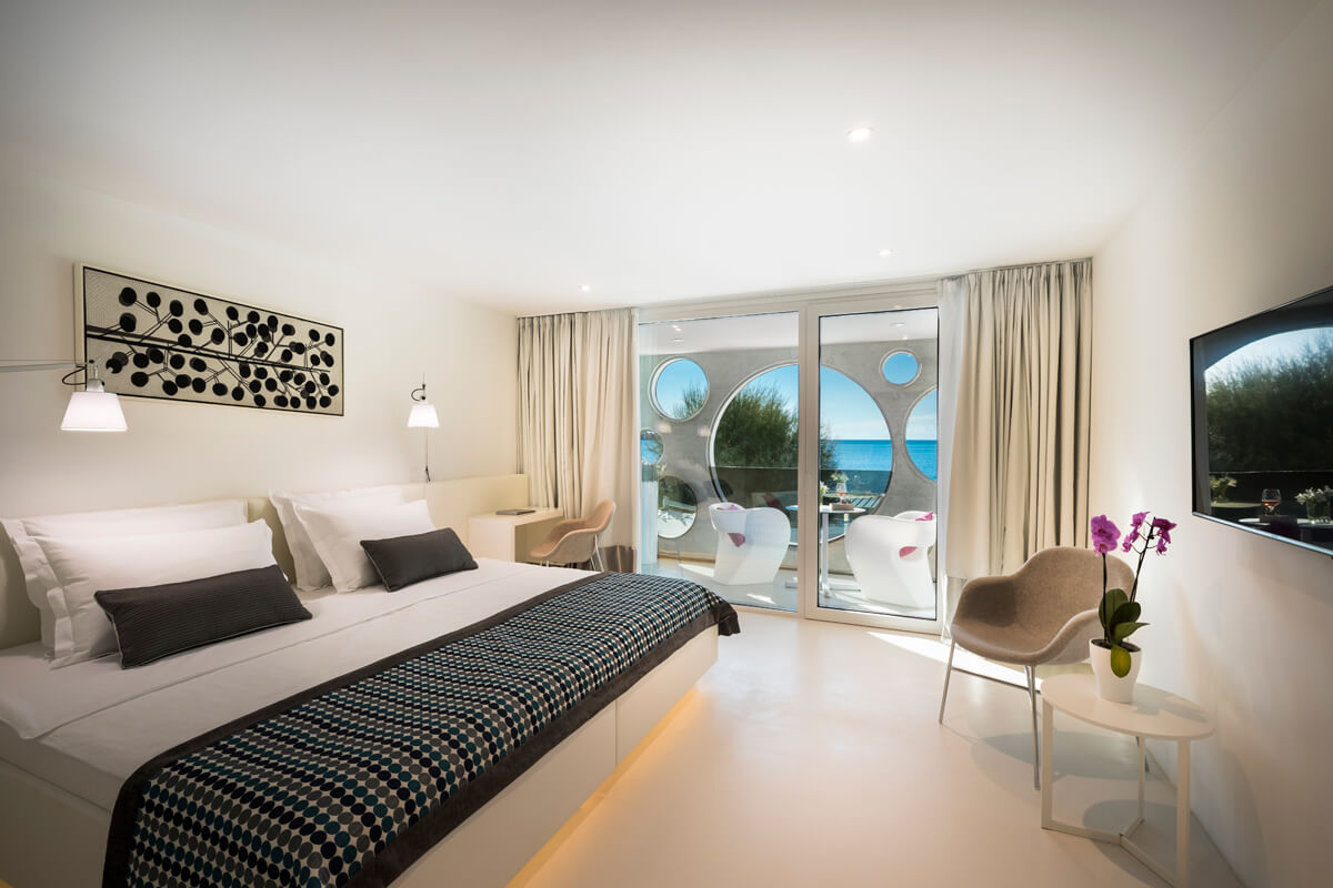 Modern eingerichtetes Hotelzimmer mit Doppelbett, großem Fenster mit Blick auf Meer, zwei Sesseln und Orchidee auf Beistelltisch