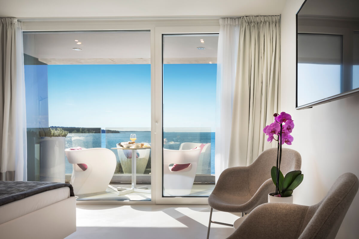 Wohnzimmer mit zwei grauen Sesseln, lila Orchidee auf Beistelltisch, Blick durch Glas-Schiebetür auf Terrasse mit weißen Stühlen und Meer im Hintergrund