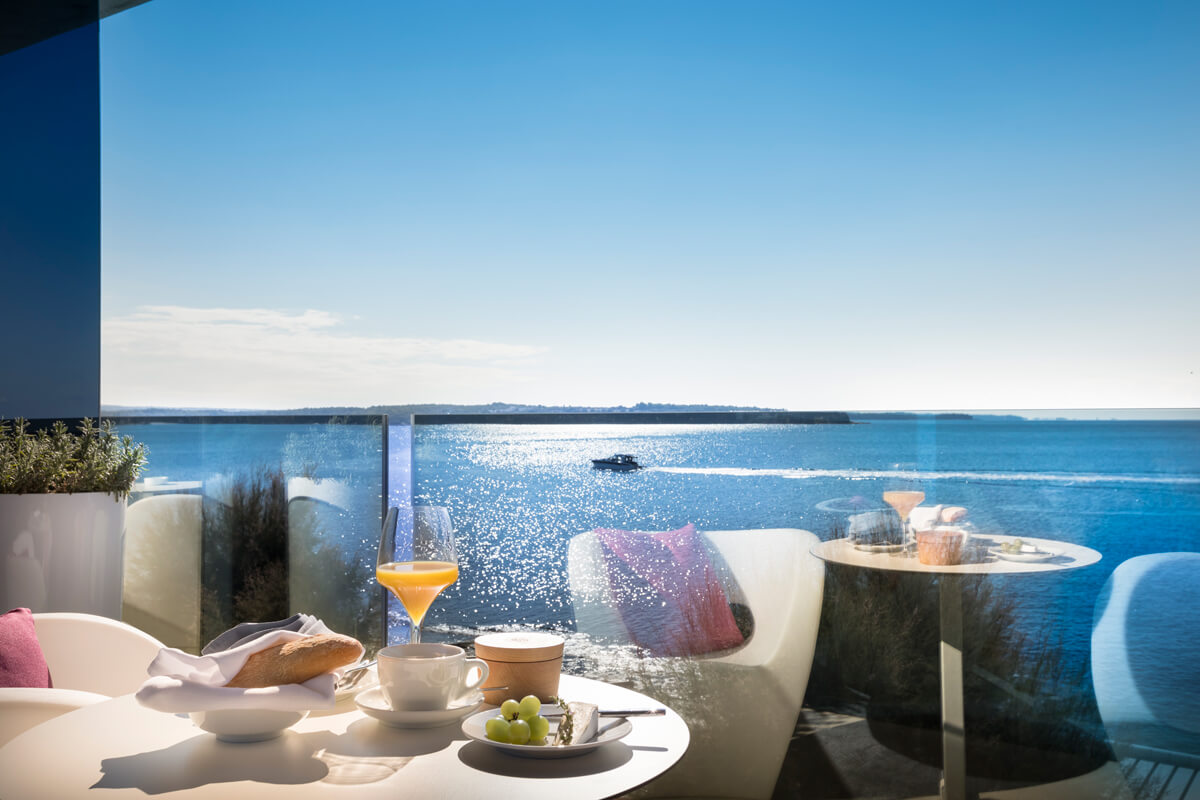 Terrasse mit gedecktem Frühstückstisch vor Meerblick, Glasgeländer und weißem Sitzmobiliar mit Kissen