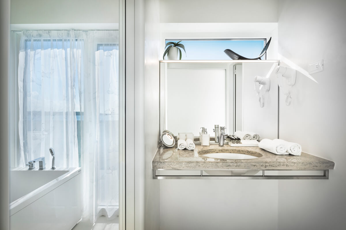 Helles Badezimmer mit Badewanne links, Glasduschwand, großem Spiegel und Waschbecken mit Marmorplatte rechts, Handtücher und Haartrockner an der Wand.