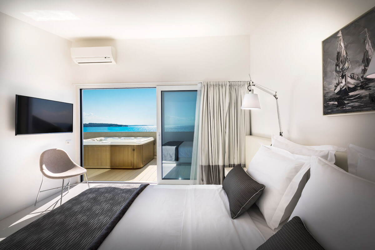 Schlafzimmer mit Doppelbett, grauen Kissen und Decke, Stuhl, Wandbild mit Segelboot, Klimaanlage und Blick auf Terrasse mit Whirlpool und Meer