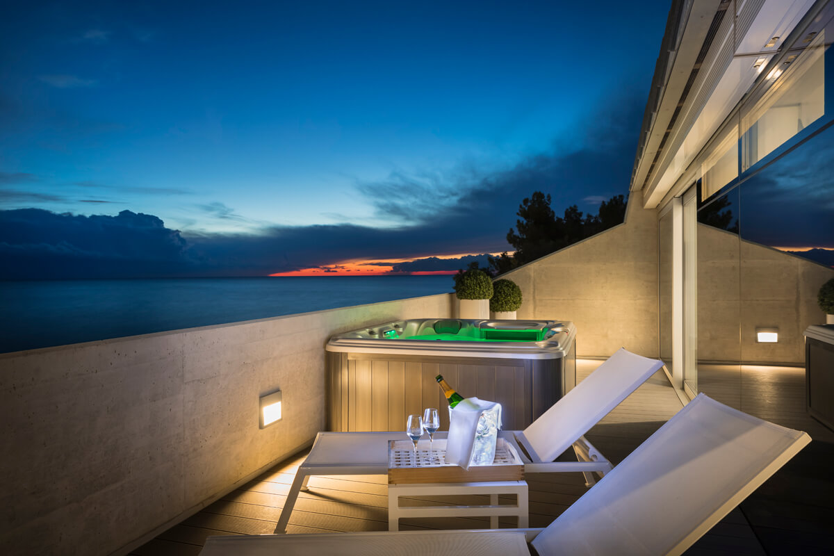 Terrasse mit zwei Liegestühlen, kleinem Tisch mit Champagnerflasche und Gläsern, Whirlpool und Meerblick bei Abenddämmerung