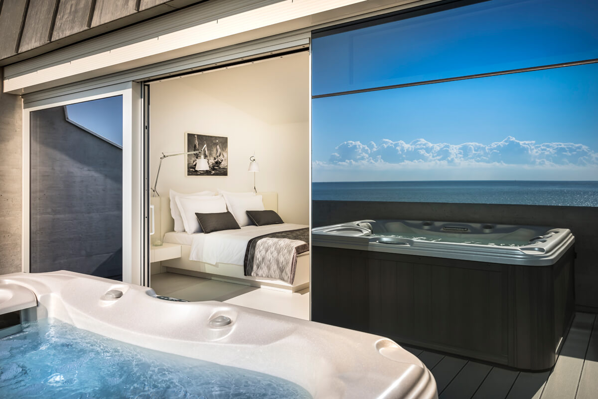 Whirlpool auf Terrasse mit Meerblick, angrenzend ein Schlafzimmer mit Doppelbett, zwei Kissen und schwarz-weißem Foto an der Wand