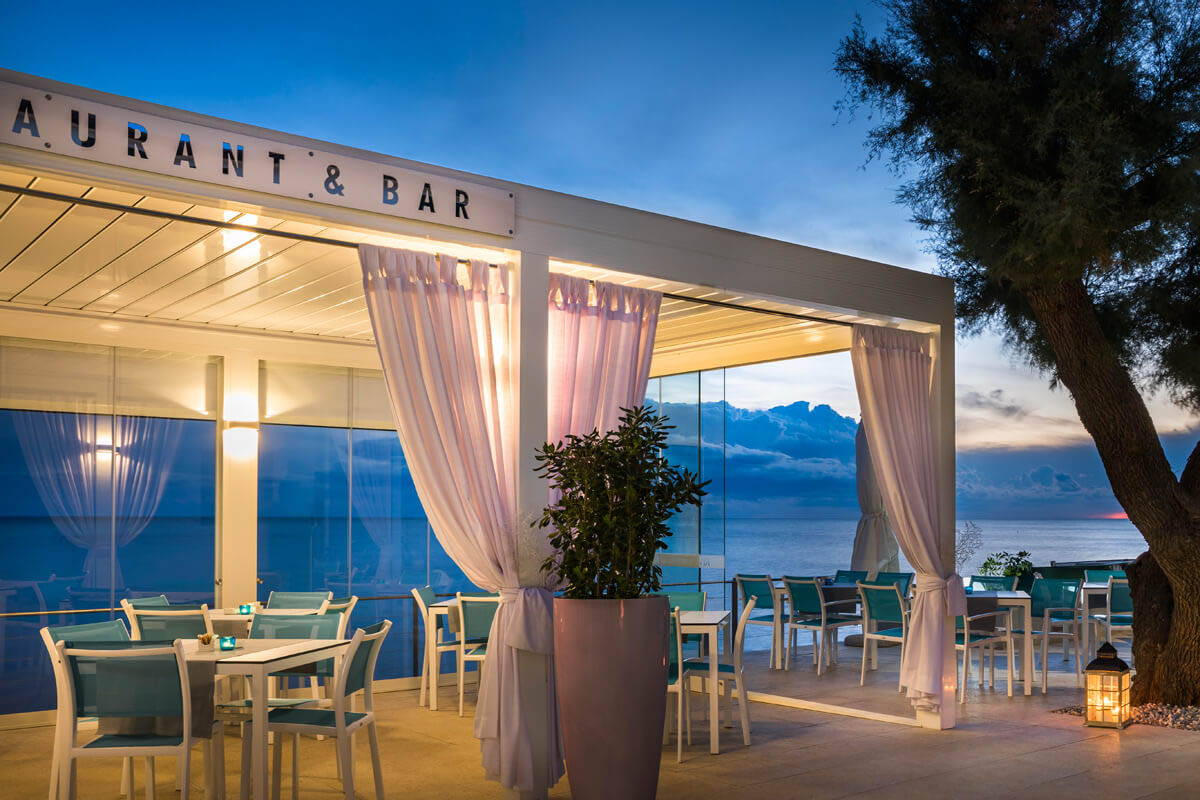 Offenes Restaurant mit weißen Tischen und Stühlen, durchsichtigen Vorhängen und Blick auf das Meer bei Abenddämmerung.