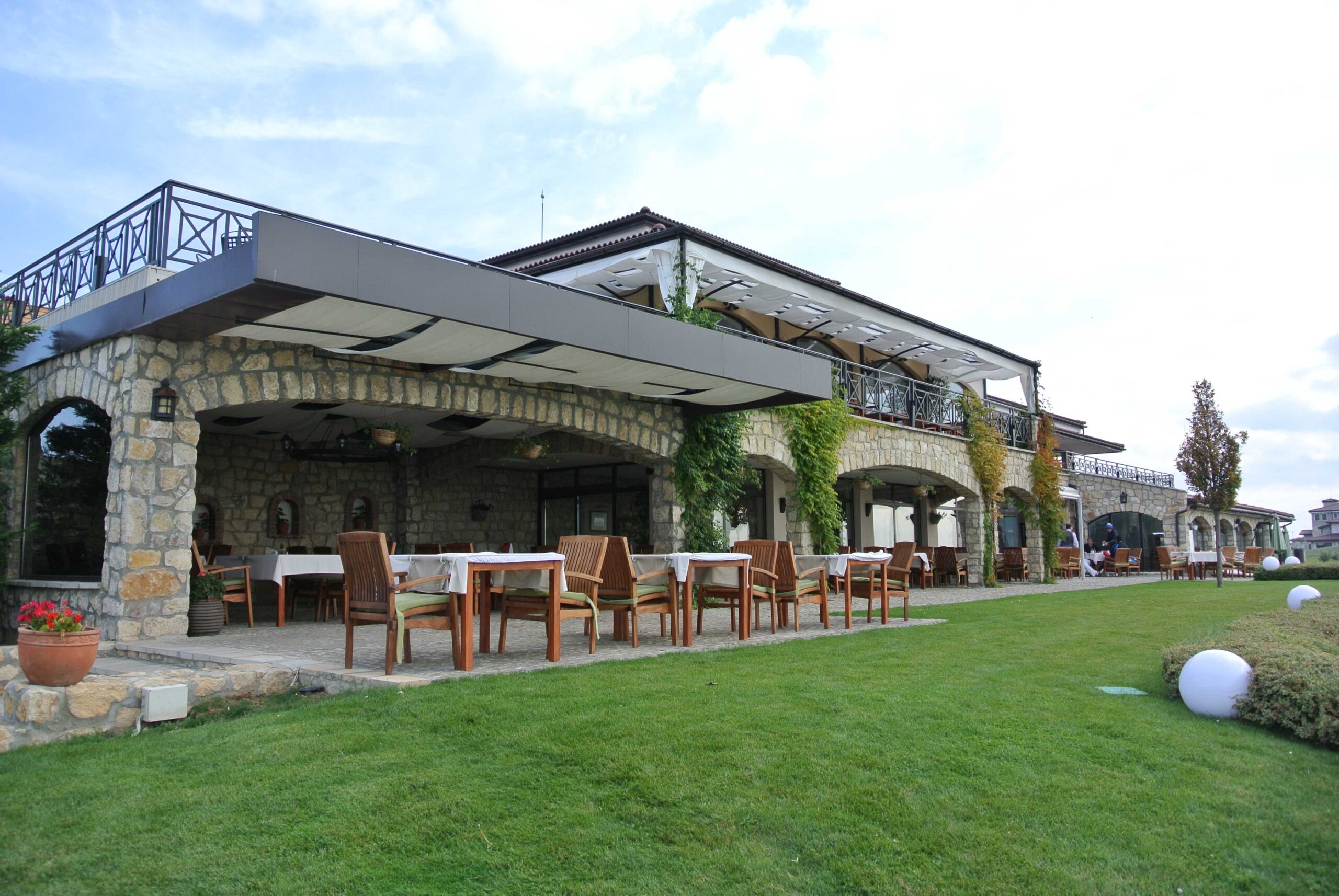Weitwinkelansicht des Resorts mit Steinbögen, überdachter Terrasse, Tischen und Stühlen im Freien sowie Rasenfläche im Vordergrund.