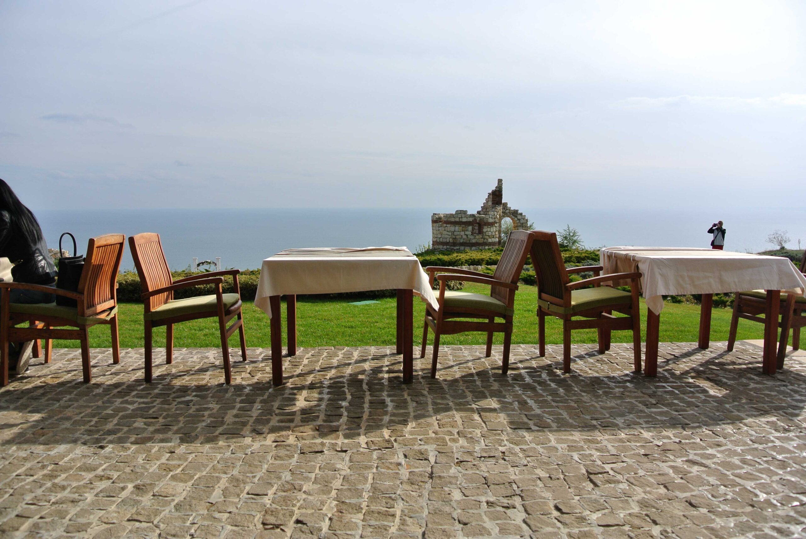 Terrasse mit Tischen und Stühlen aus Holz, Blick auf eine historische Steinruine und das Meer im Hintergrund.