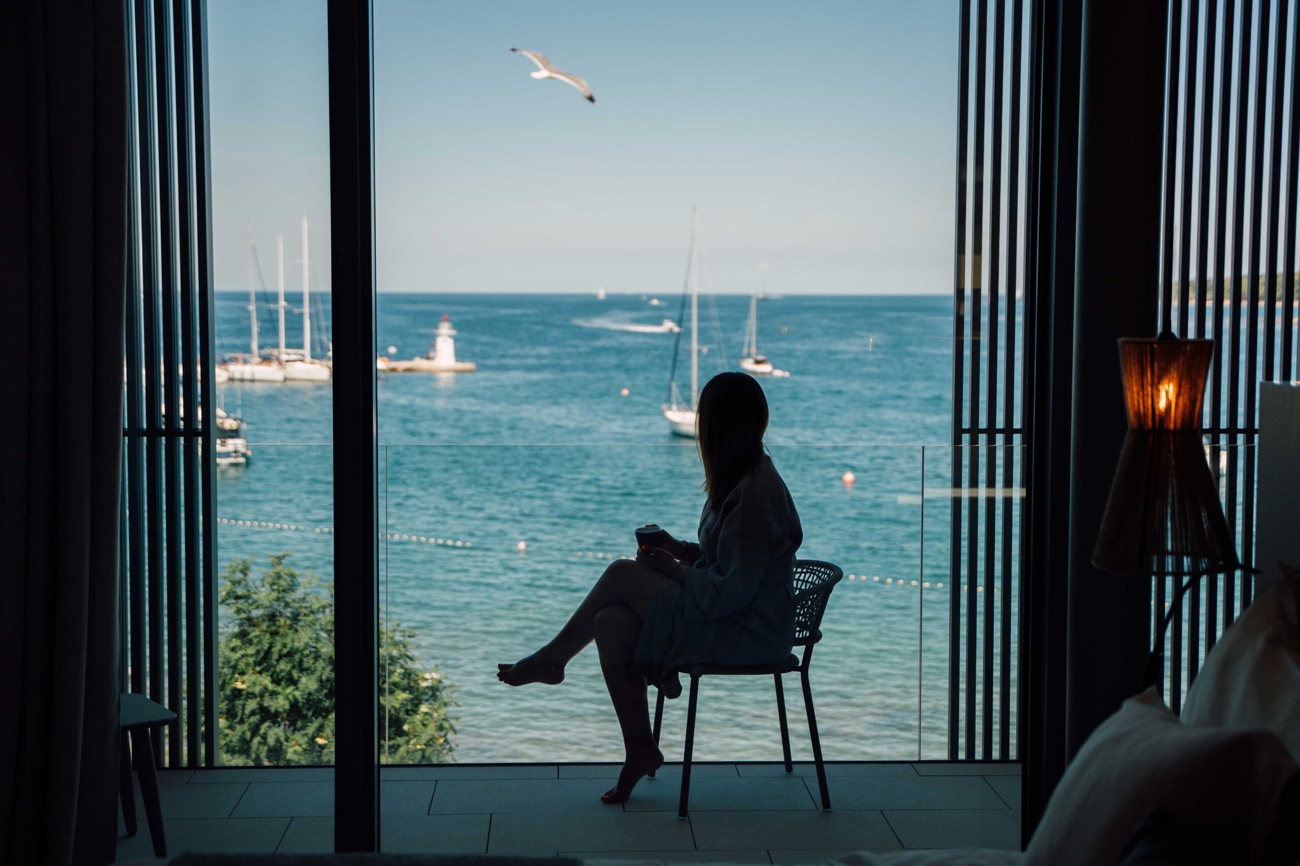 Silhouette einer Frau, die auf einem Stuhl vor einer großen Fensterfront sitzt, mit Blick auf das Meer, Segelboote und einen Leuchtturm.