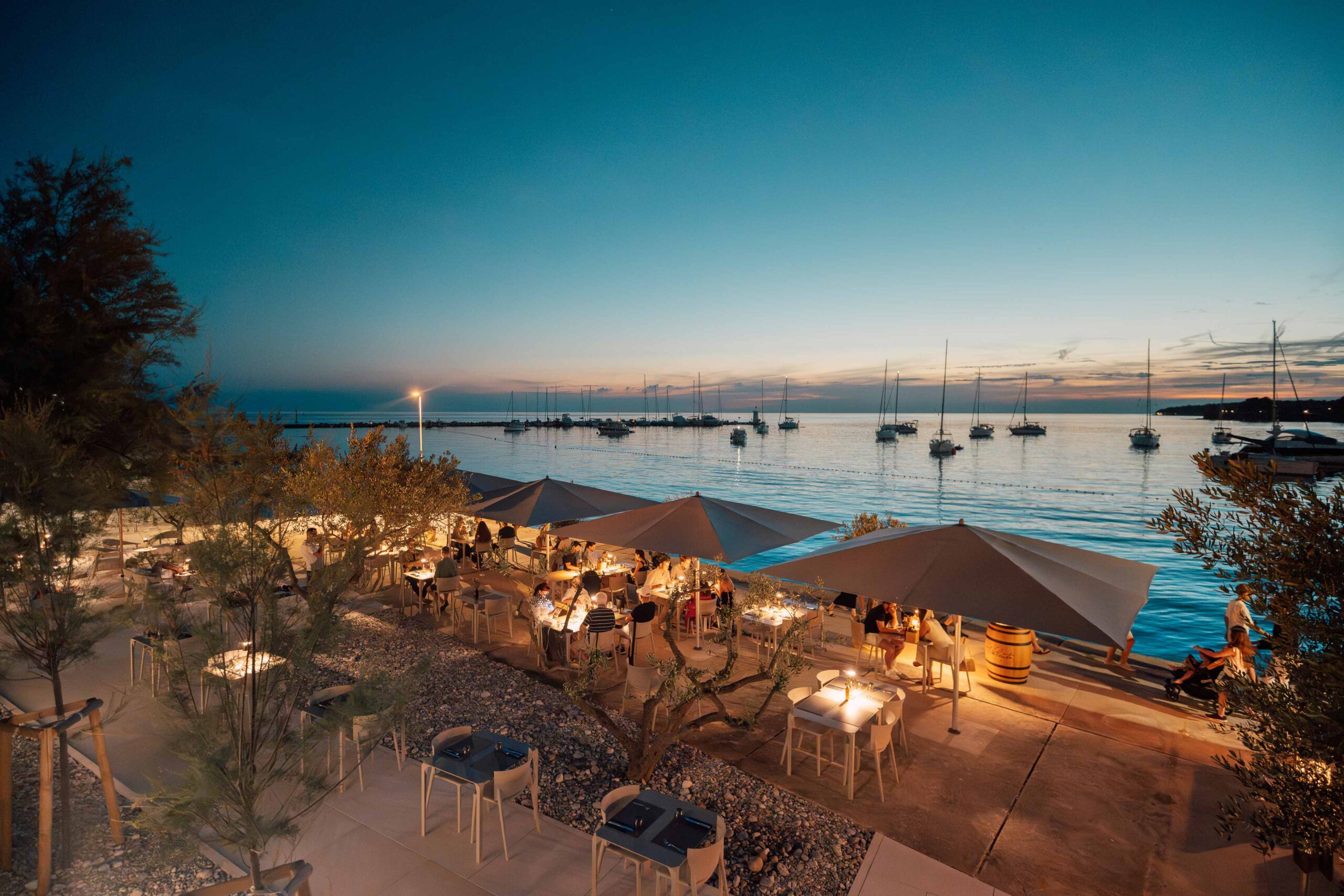 Abendstimmung an einer Restaurantterrasse direkt am Meer mit beleuchteten Tischen und Blick auf Segelboote im Wasser.