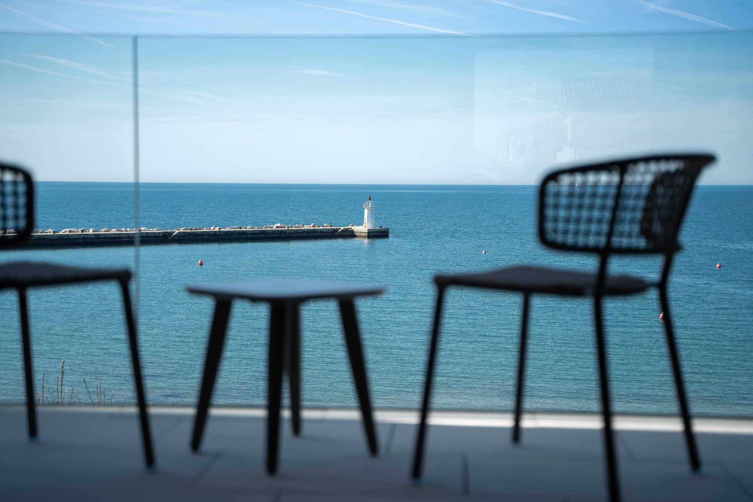Blick durch Glasbalkon auf einen kleinen Leuchtturm am Ende einer Mole, eingerahmt von zwei unscharfen Stühlen im Vordergrund.