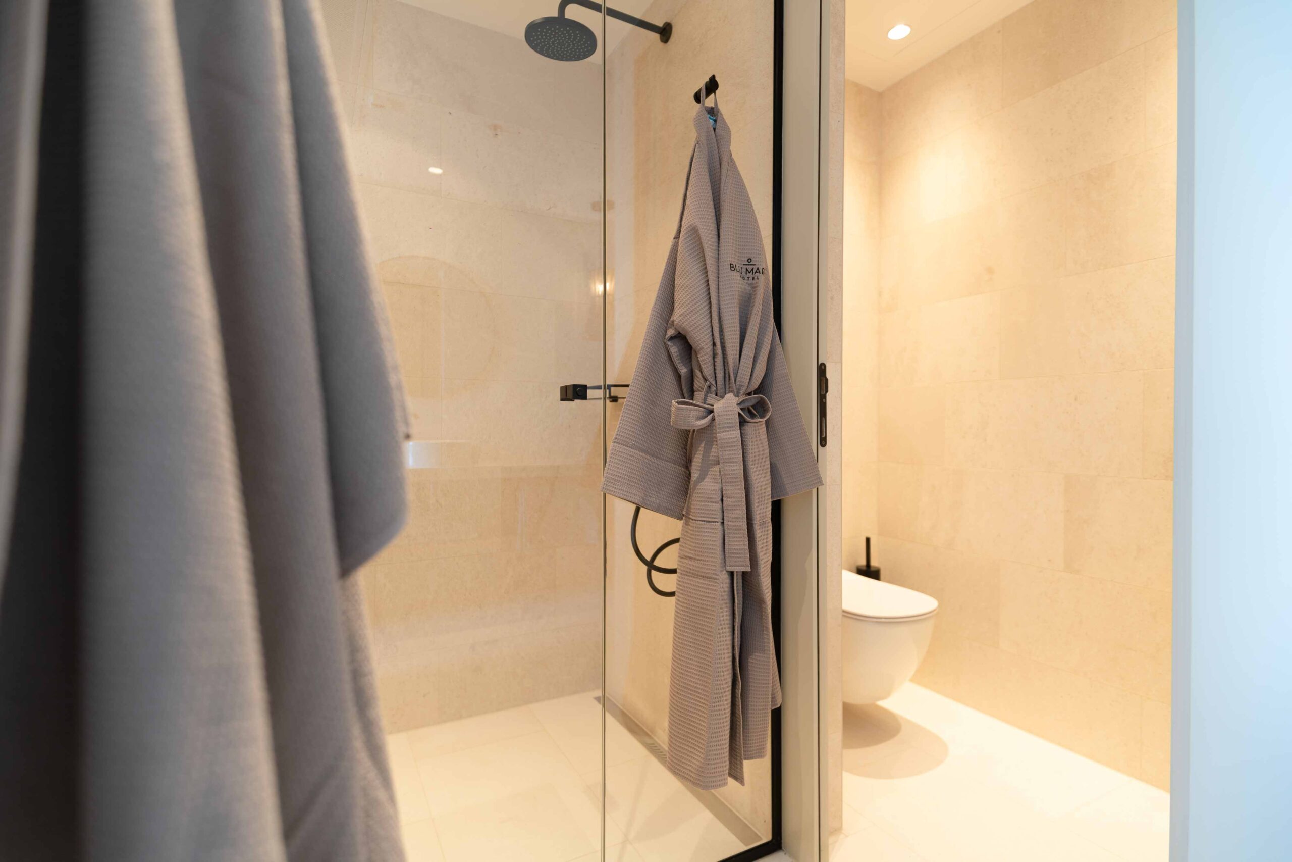 Modernes Badezimmer mit Regendusche, beigefarbenen Fliesen und einem an der Tür hängenden Bademantel.