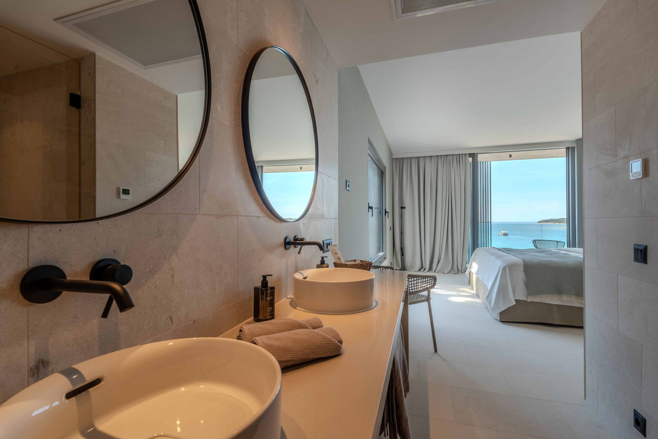 Helles Badezimmer mit zwei runden Spiegeln, Doppelwaschbecken und Blick in ein Schlafzimmer mit Meerblick.