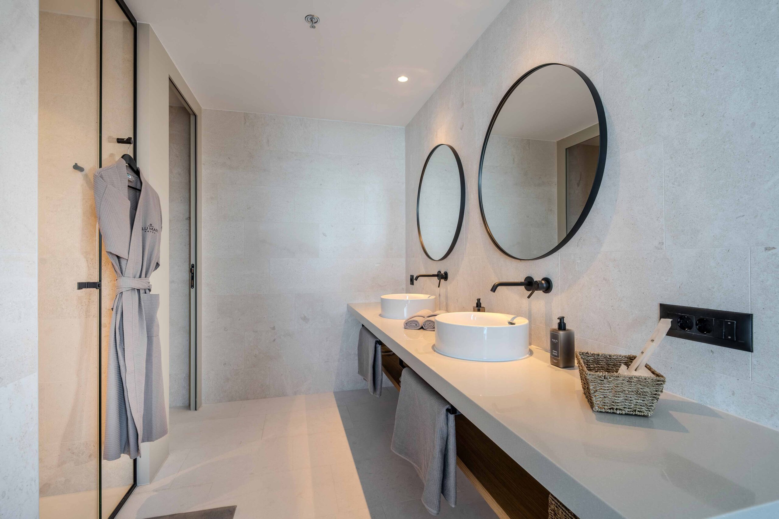 Modernes Badezimmer mit Doppelwaschbecken, zwei runden Spiegeln und einem Bademantel an der Duschkabinentür.