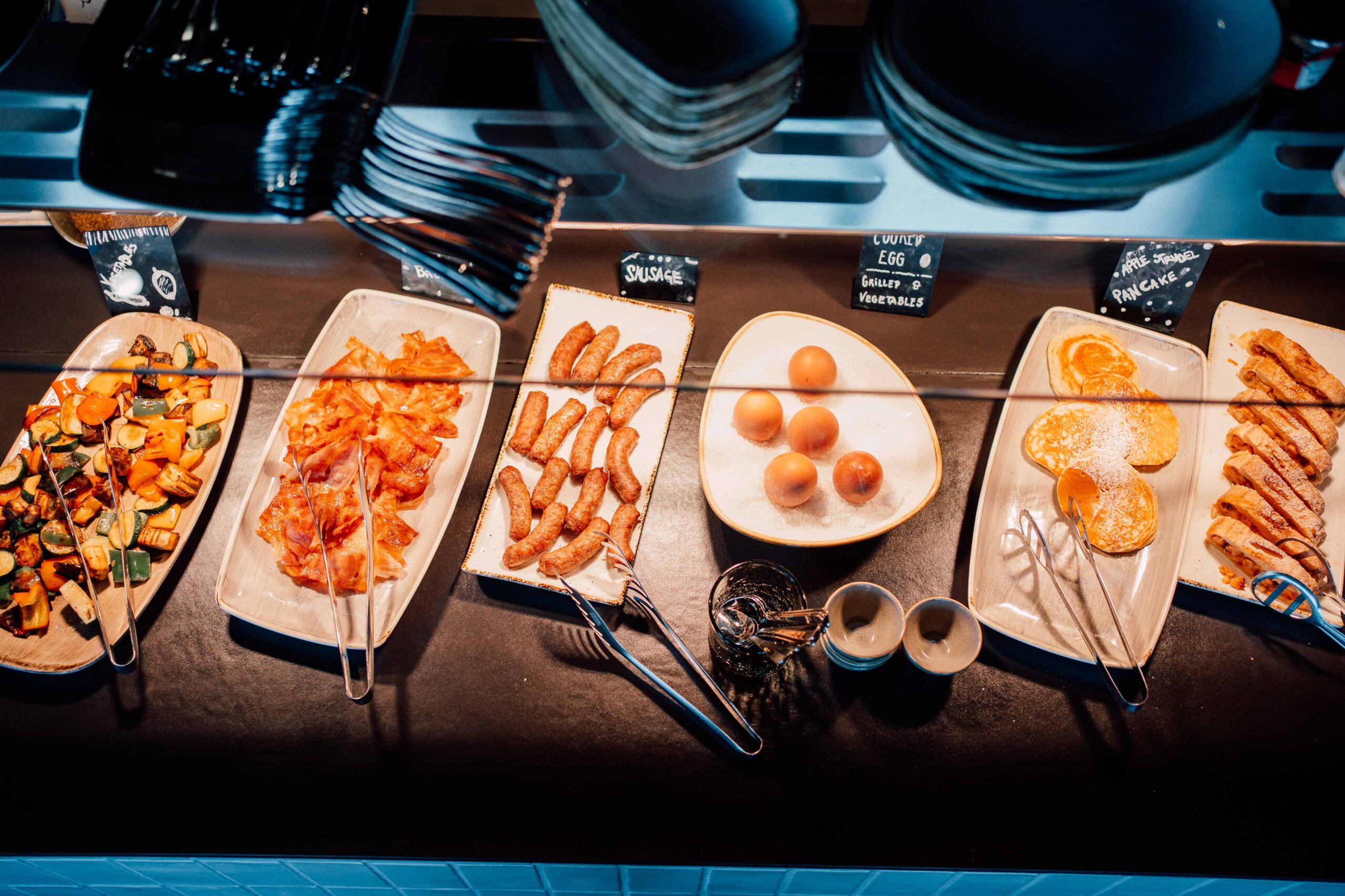 Warmes Frühstücksbuffet mit gegrilltem Gemüse, Speck, Würstchen, Eiern, Pancakes und Gebäck.