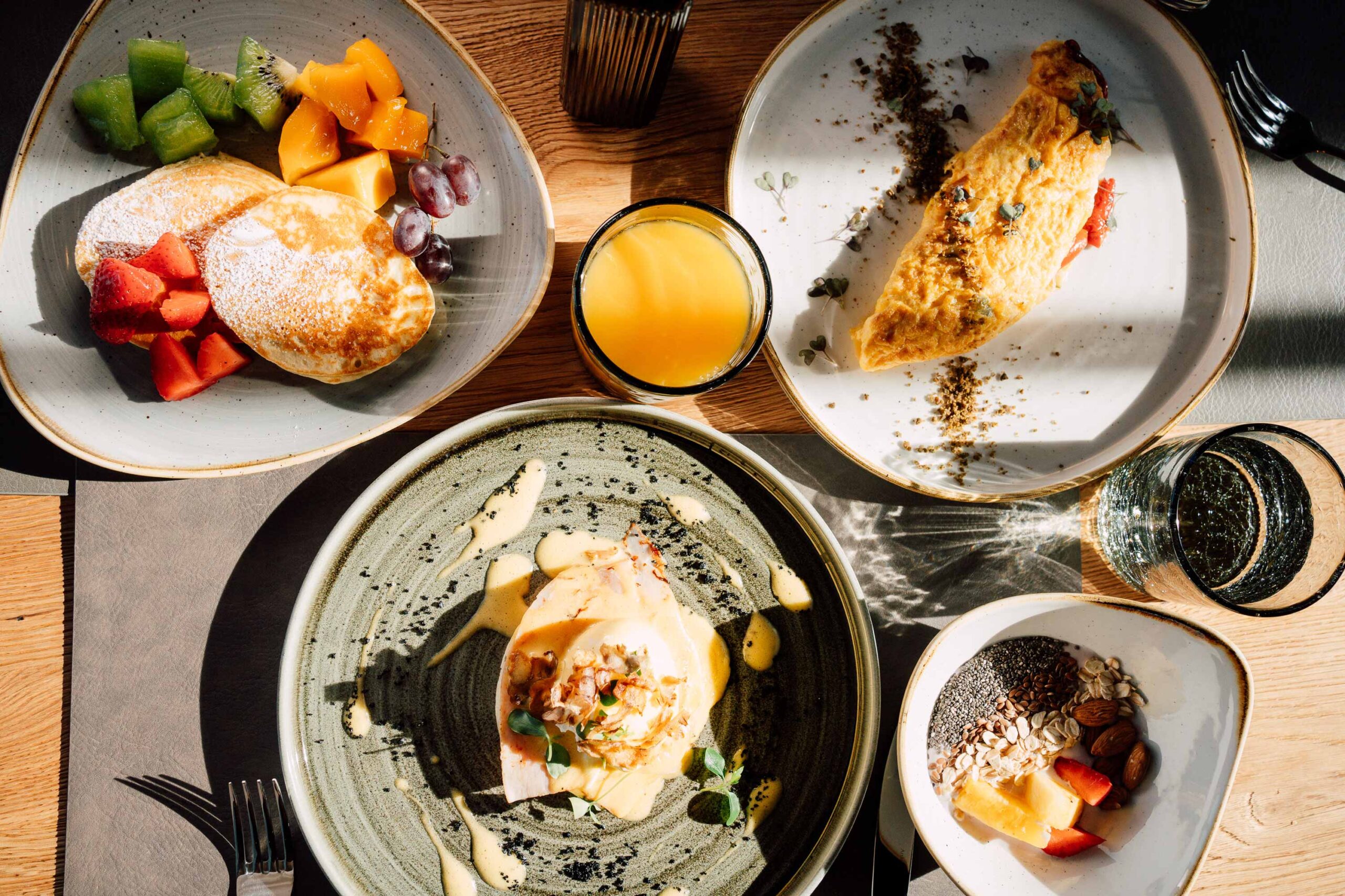 Frühstückstisch mit Pancakes und Obst, Omelette, Eggs Benedict, Müsli und einem Glas Orangensaft.