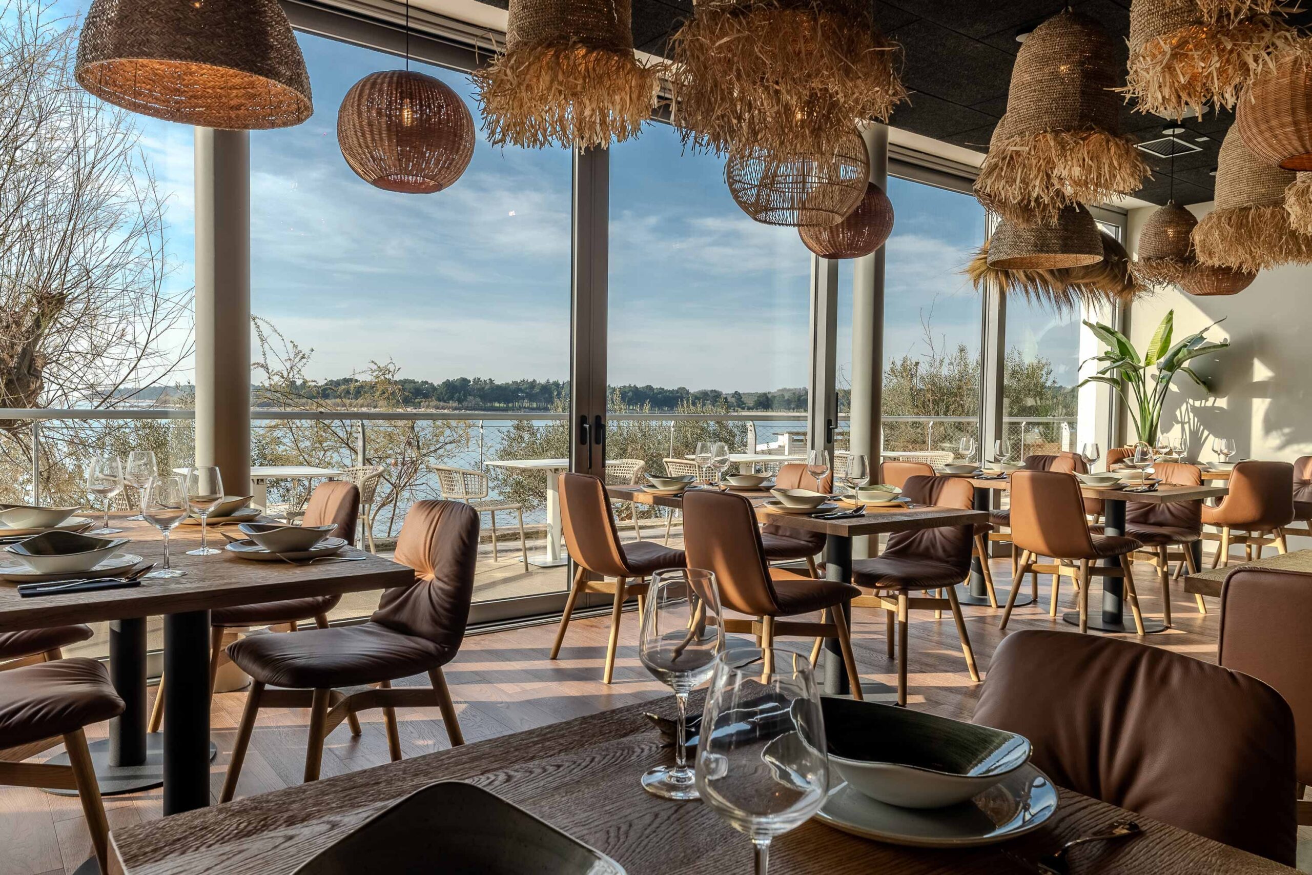 Modern eingerichtetes Restaurant mit großen Panoramafenstern, Blick auf das Wasser und warmem Tageslicht, dekoriert mit Hängelampen aus Naturmaterialien.