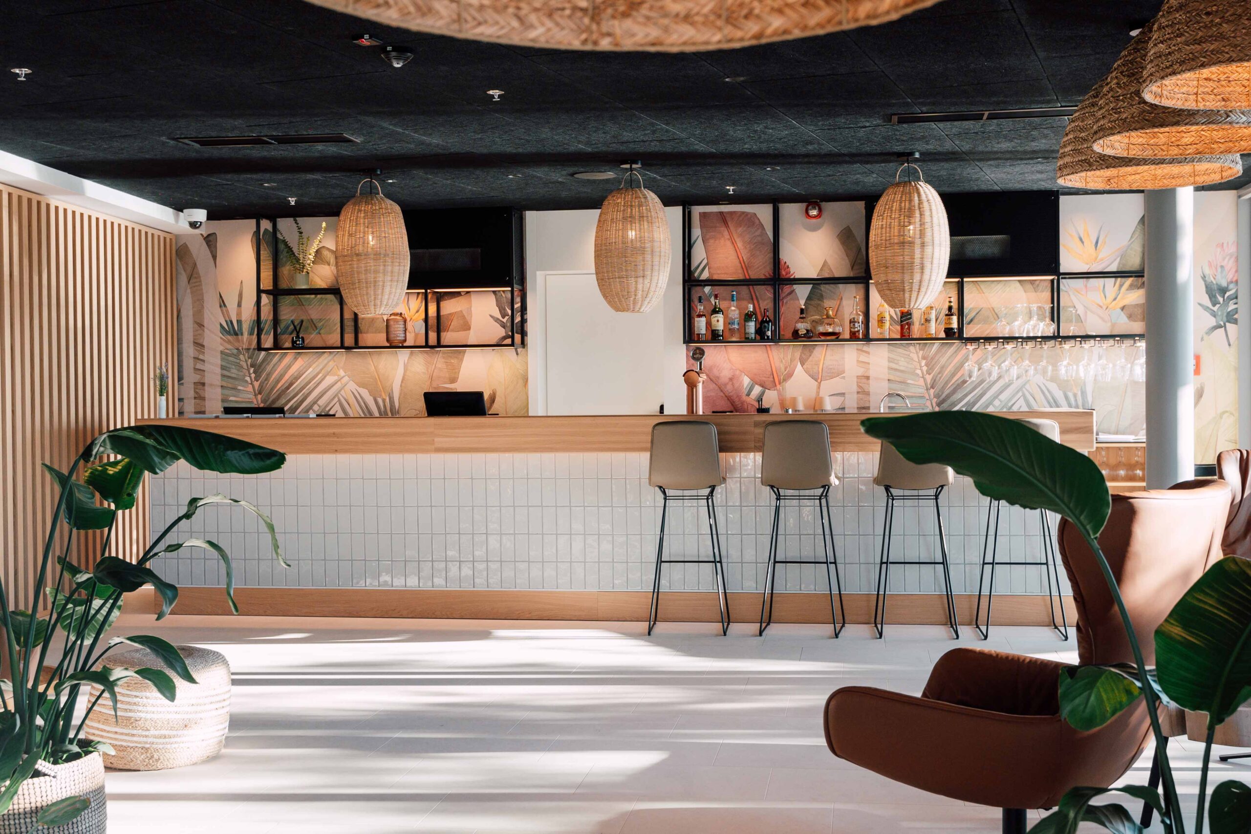 Stilvolle Hotelbar mit Holztresen, hohen Barhockern, Regalen mit Flaschen und dekorativer Wandgestaltung mit tropischen Pflanzenmotiven.