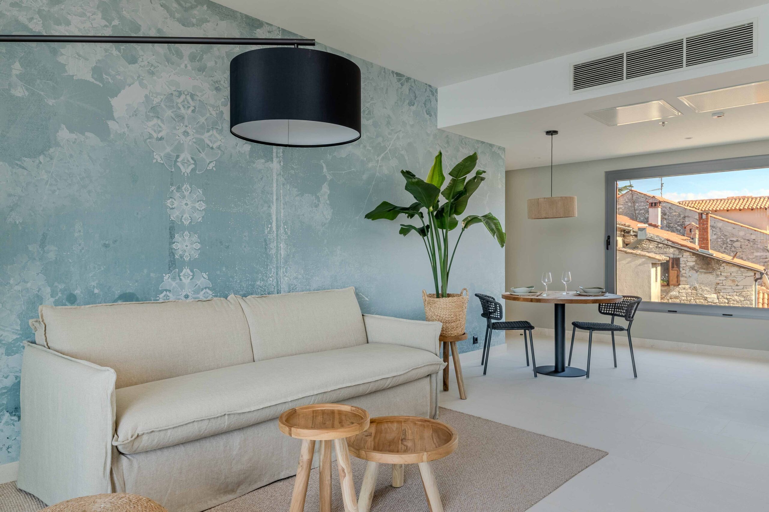 Helles Wohnzimmer mit beiger Couch, hellblauer Strukturwand, runden Holztischen und Esstisch mit Blick auf ein mediterranes Dorf.