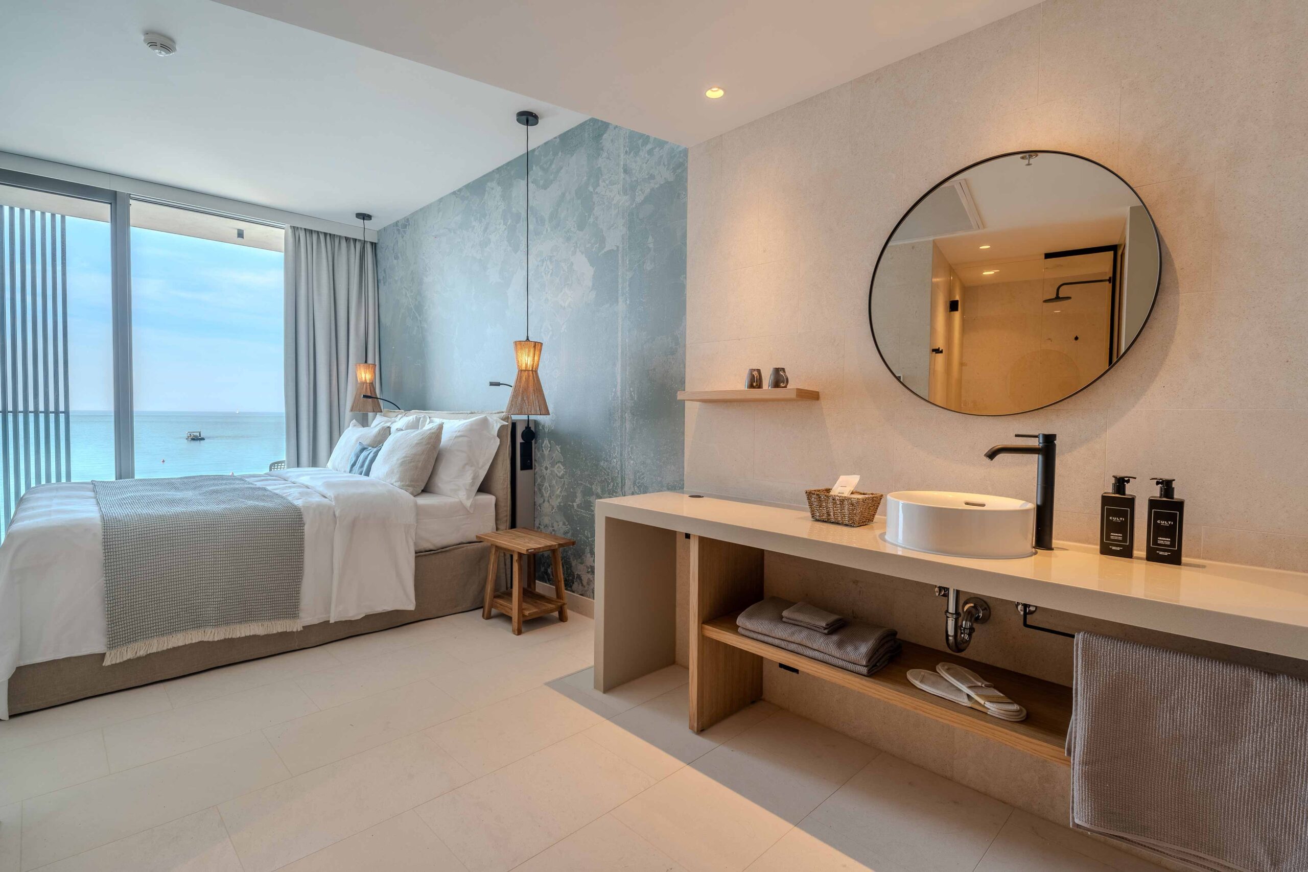 Hotelzimmer mit großem Doppelbett, Meerblick, integrierter Waschtisch- und Badbereich mit rundem Spiegel und modernen Armaturen.