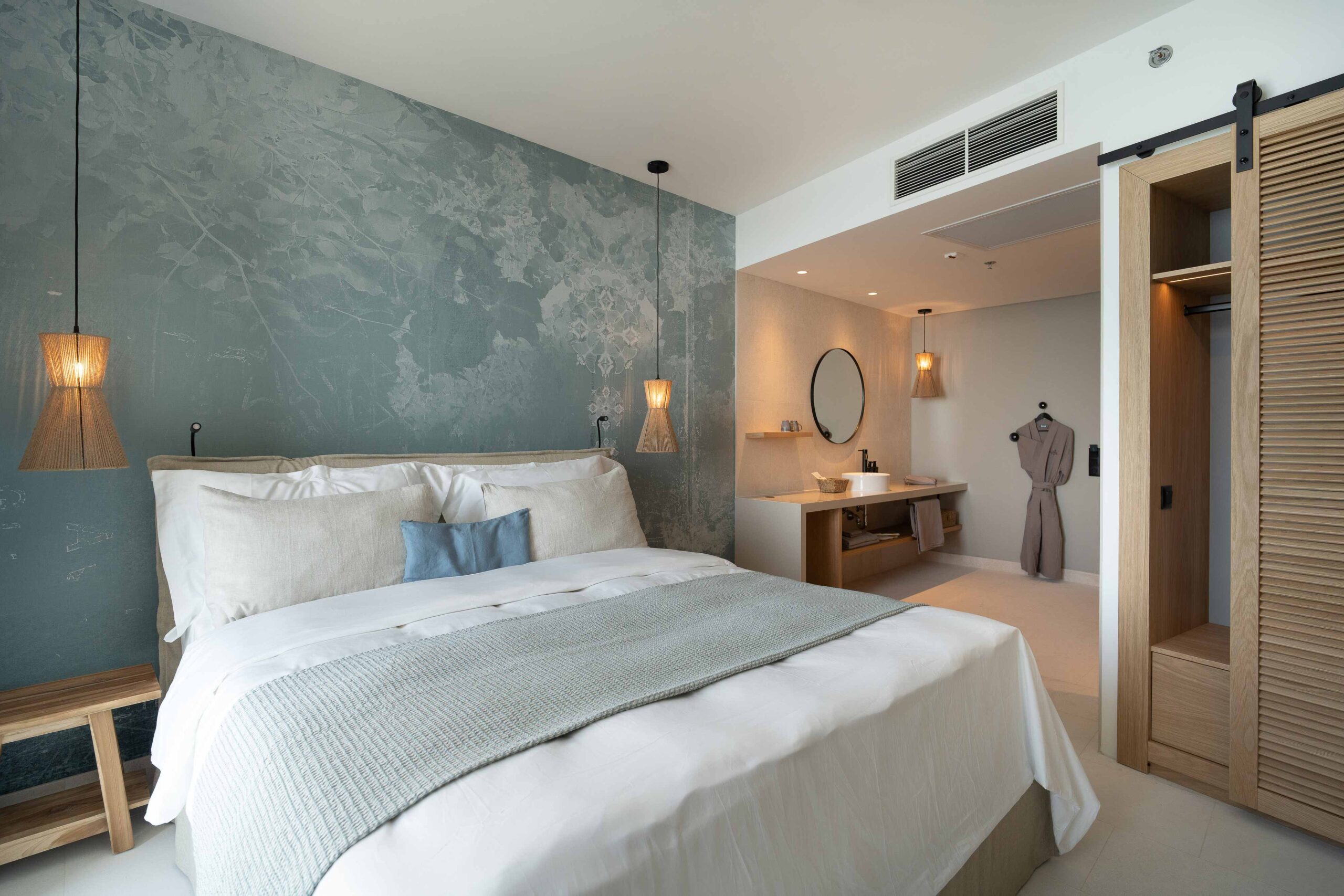 Hotelzimmer mit großem Doppelbett, Blick auf Bad- und Ankleidebereich, hellblauer Akzentwand und warmen Holzdetails.