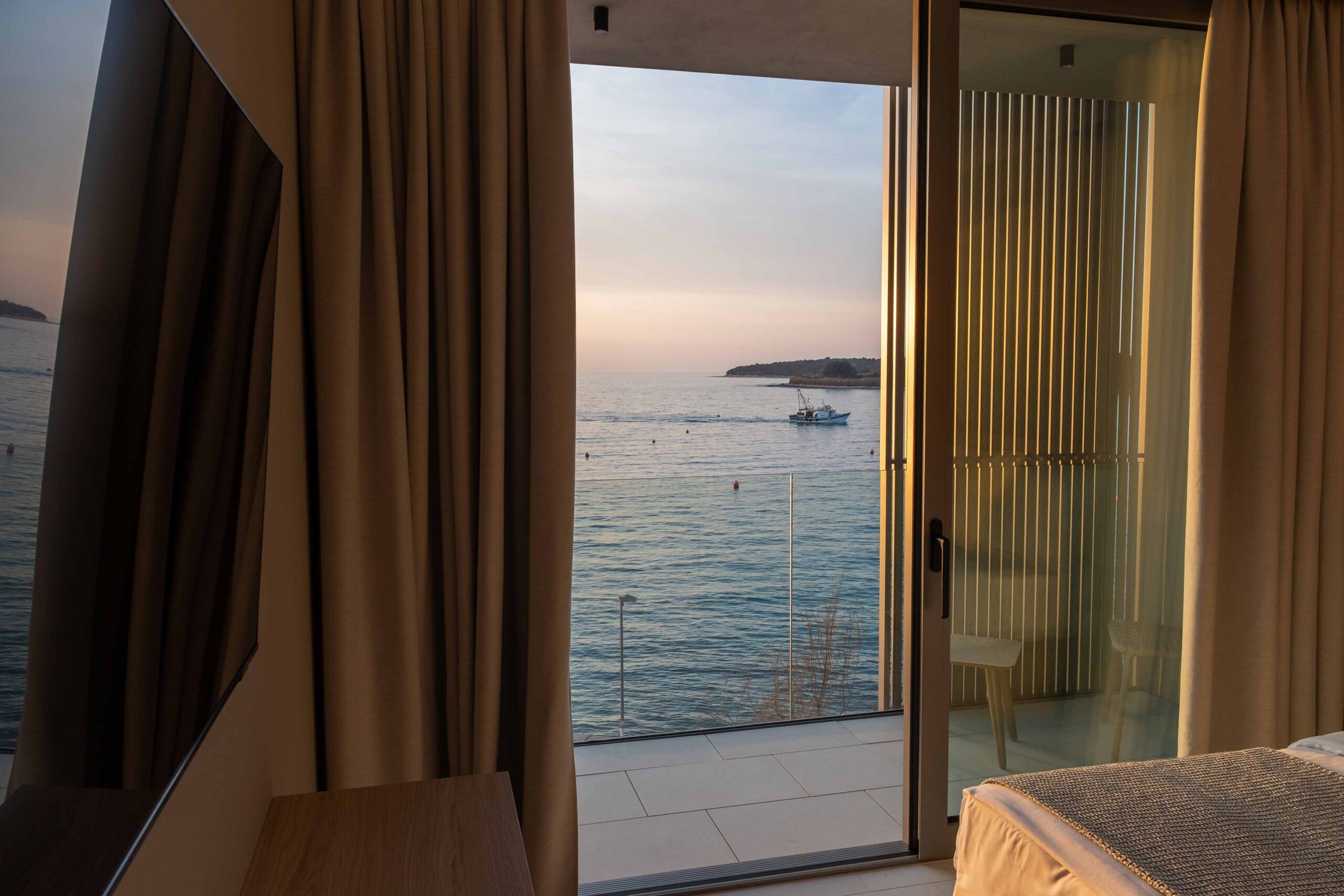 Blick aus einem Hotelzimmer durch eine offene Glastür auf das Meer mit einem Fischerboot bei Sonnenuntergang.