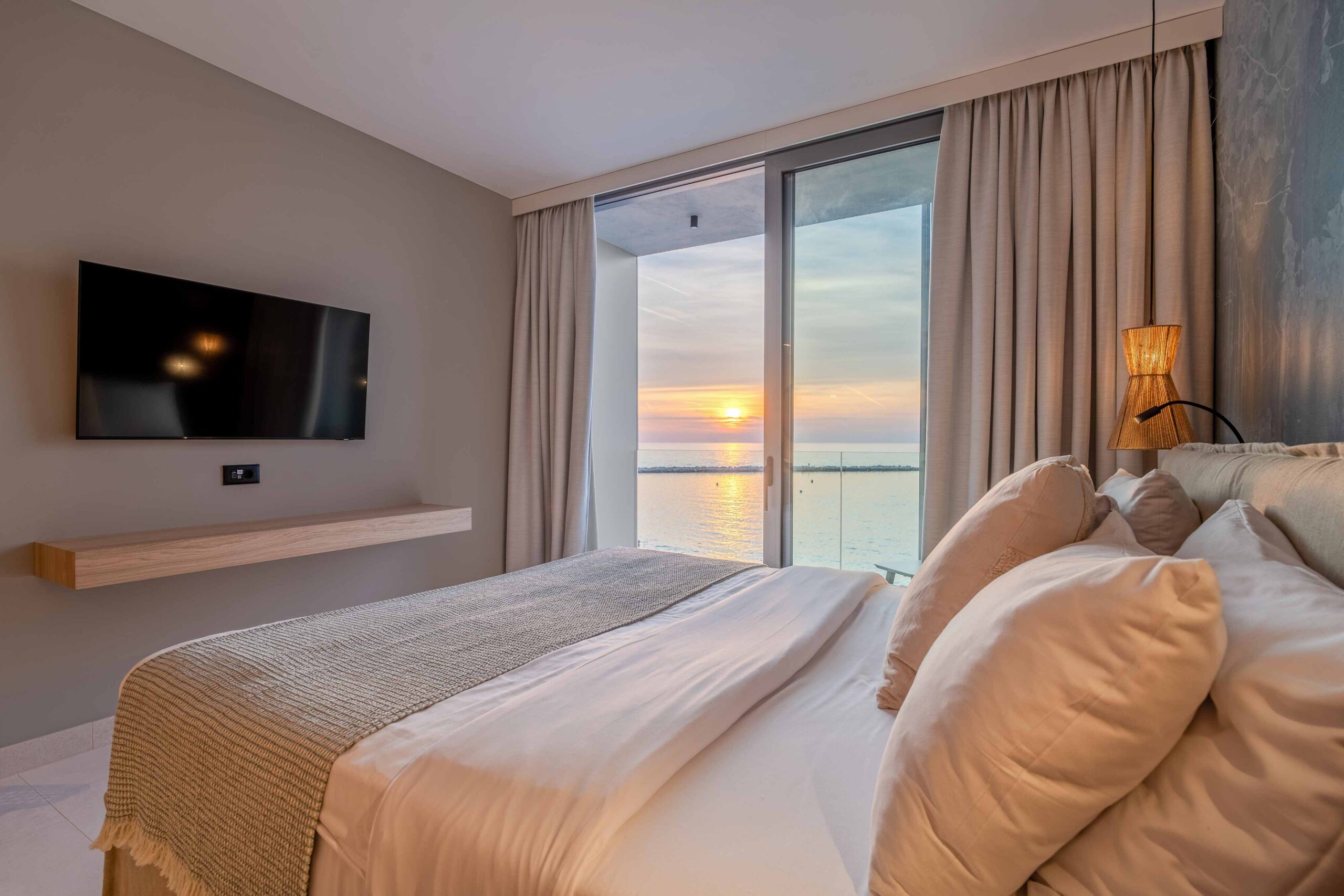 Hotelzimmer mit Doppelbett, Blick durch bodentiefe Fenster auf einen Sonnenuntergang über dem Meer, Fernseher an der Wand.