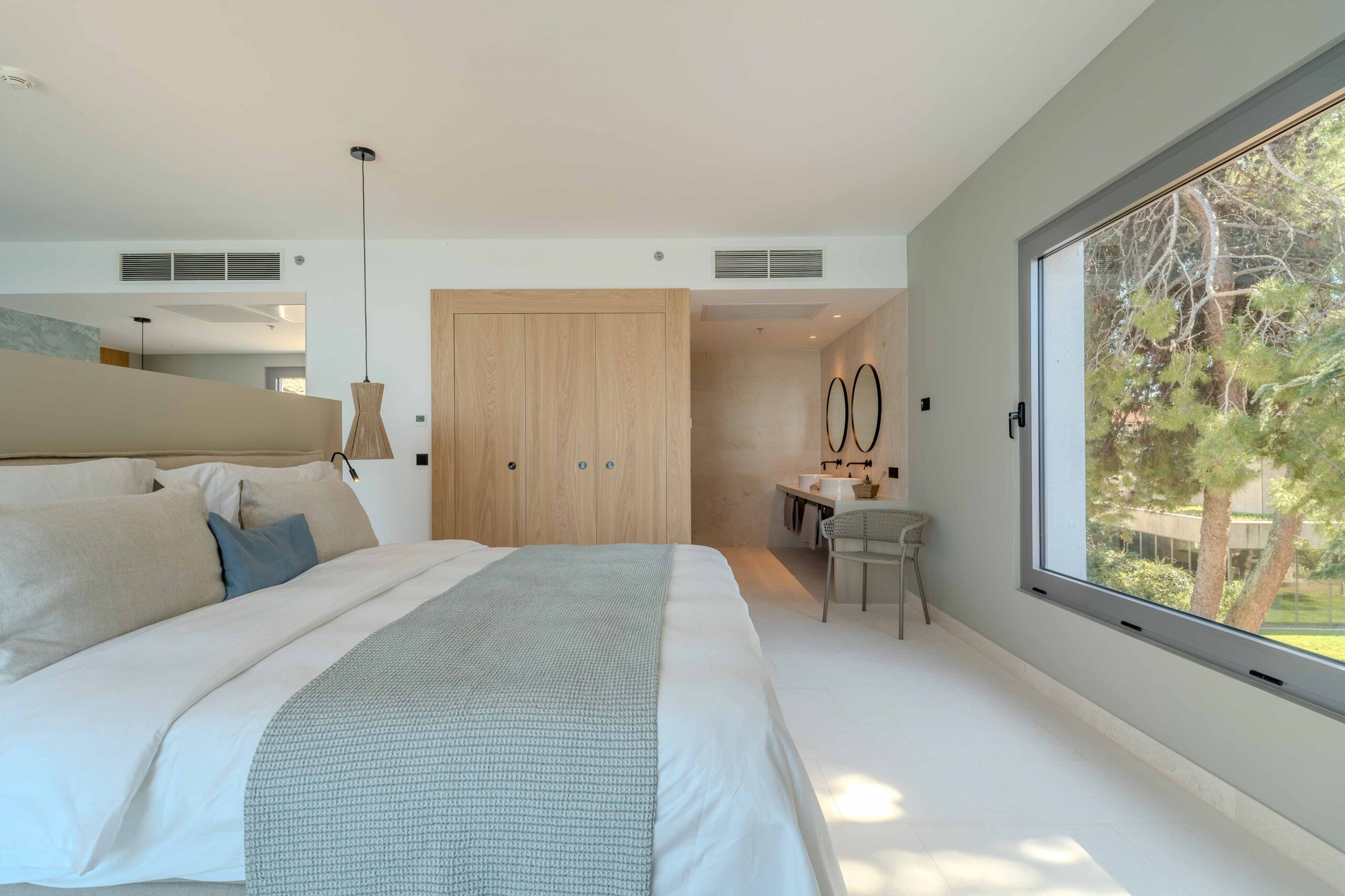 Geräumiges Hotelzimmer mit großem Doppelbett, hellem Interieur, Blick ins Badezimmer mit zwei runden Spiegeln, Fenster mit Gartenblick.
