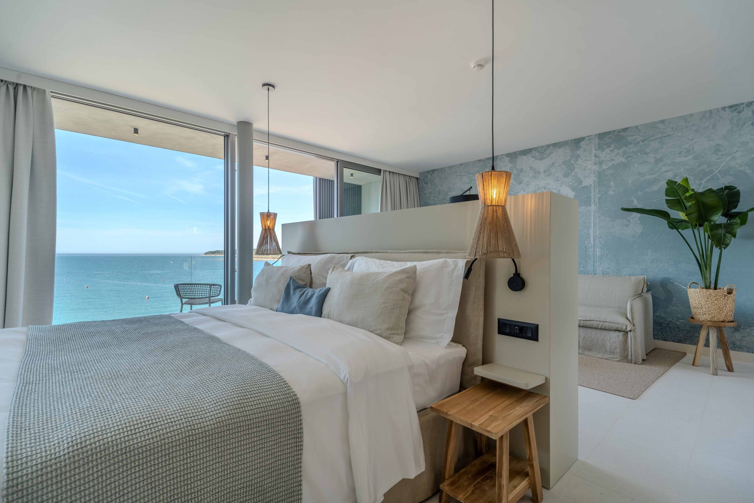 Helles Hotelzimmer mit Doppelbett, blau gemusterter Wand, Meerblick durch bodentiefe Fenster und Sitzecke im Hintergrund.