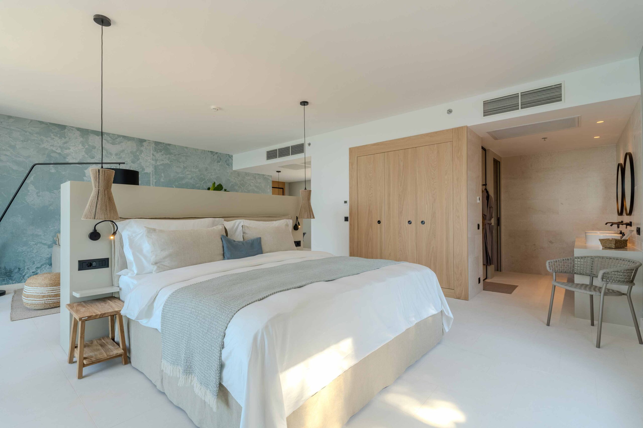 Modernes Hotelzimmer mit großem Doppelbett, Holzschrank, Blick ins Badezimmer mit Doppelwaschbecken und Gartenblick durch ein großes Fenster.