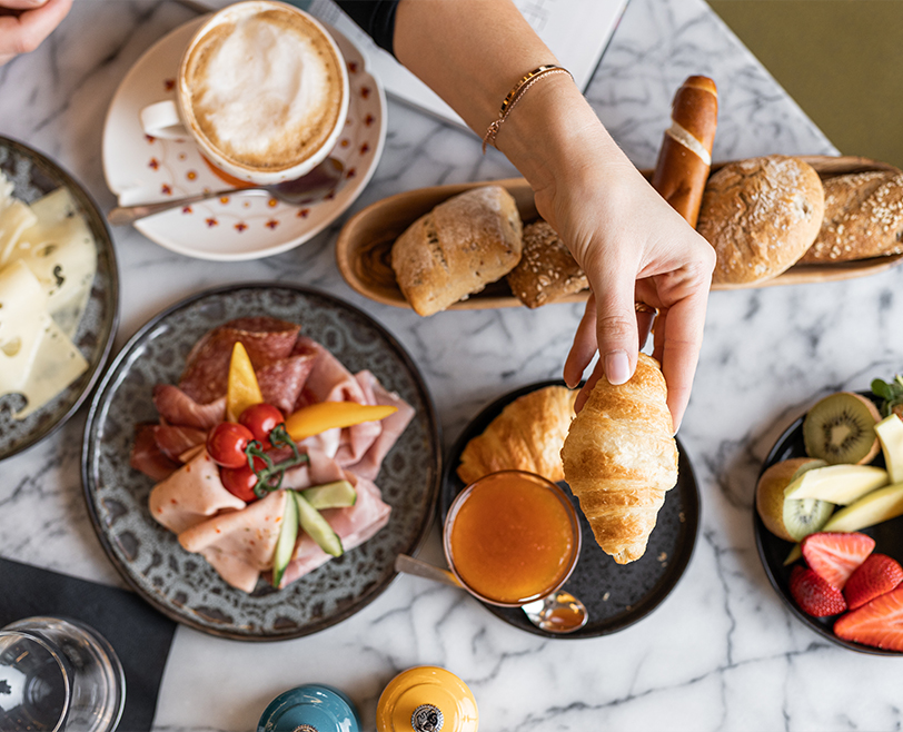 Frühstückstisch mit Croissant, frischen Brötchen, Aufschnittplatte, Obst, Kaffee und Marmelade auf Marmorplatte.