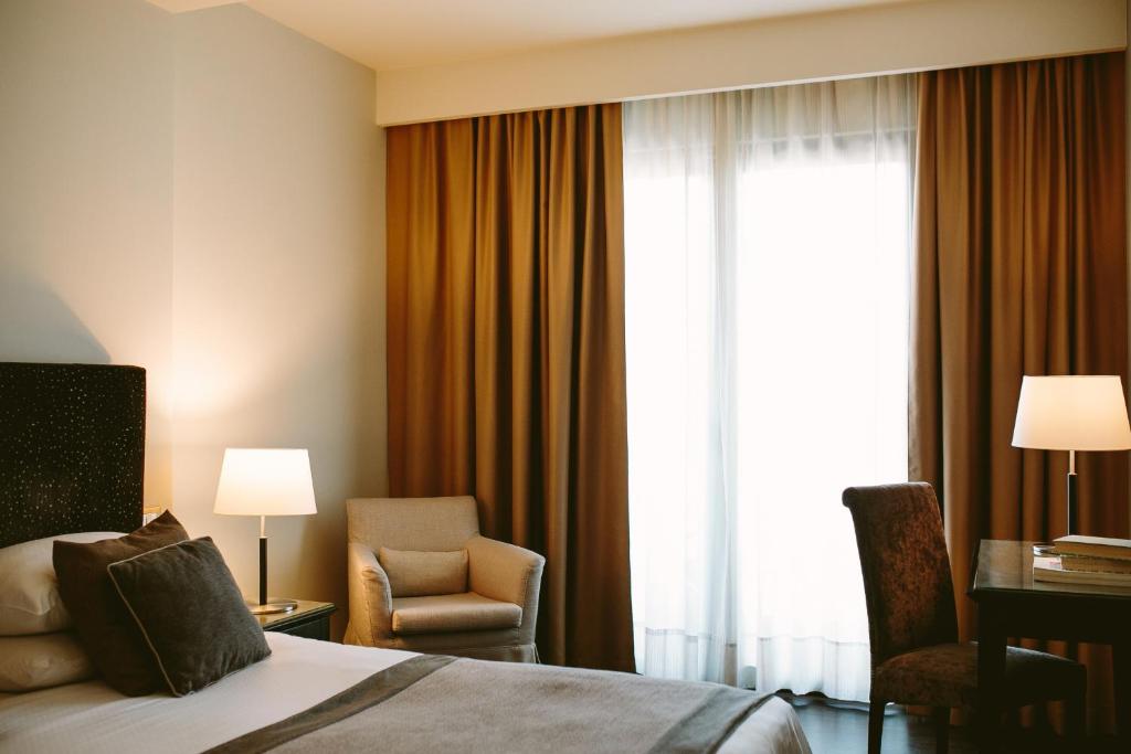 Helles Hotelzimmer mit Doppelbett, Sessel, Schreibtisch und zwei Tischlampen, großes Fenster mit braunen Vorhängen.
