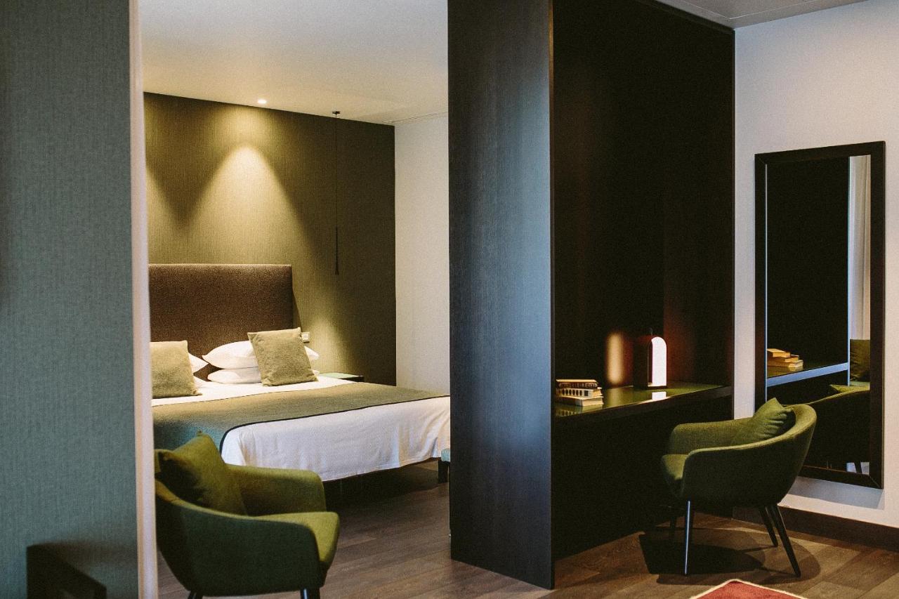 Modernes Hotelzimmer mit Doppelbett, zwei grünen Sesseln, Schreibtisch, Spiegel und stimmungsvoller Beleuchtung.