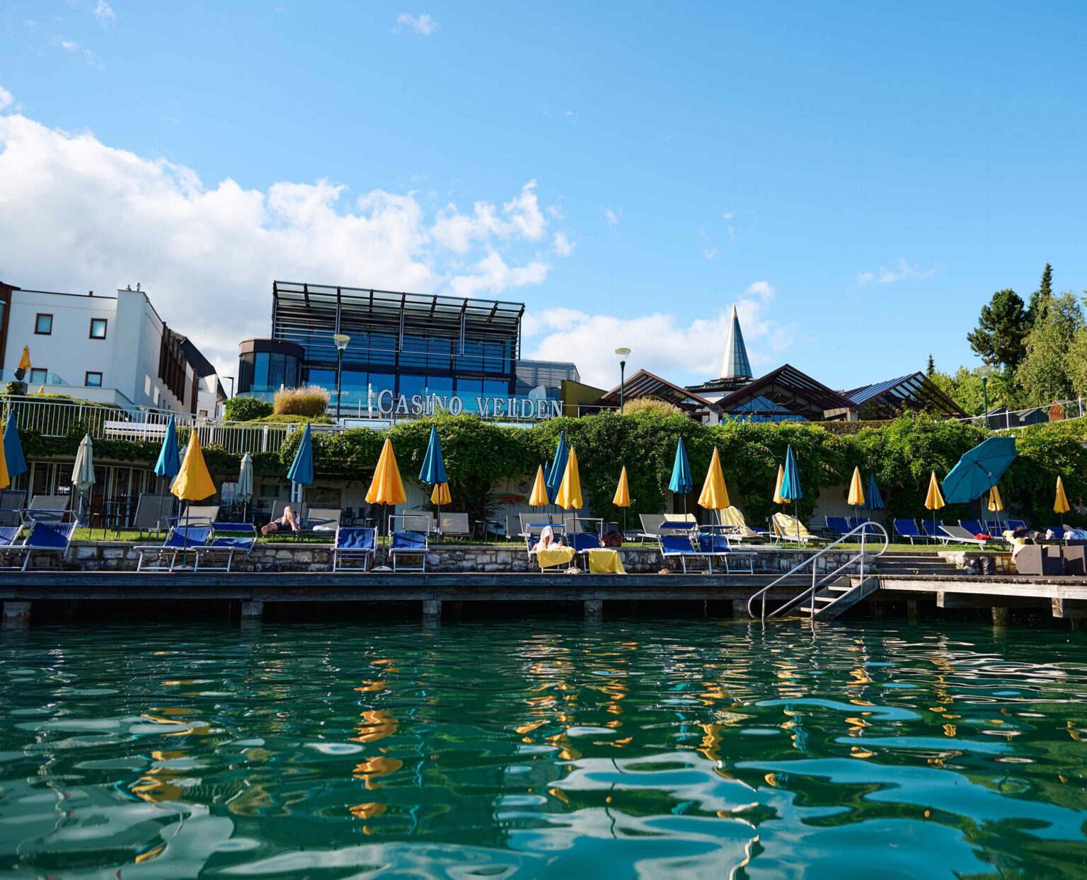 Blick vom Wasser auf das Casino Velden mit Liegewiese und bunten Sonnenschirmen am Ufer des Wörthersees.