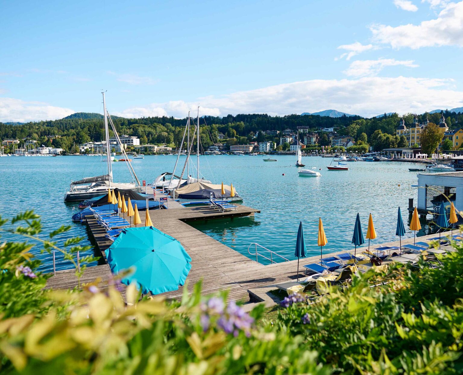 Steg mit Sonnenliegen und Sonnenschirmen am Wörthersee, Segelboote und Yachten im Wasser, Blick auf Velden.