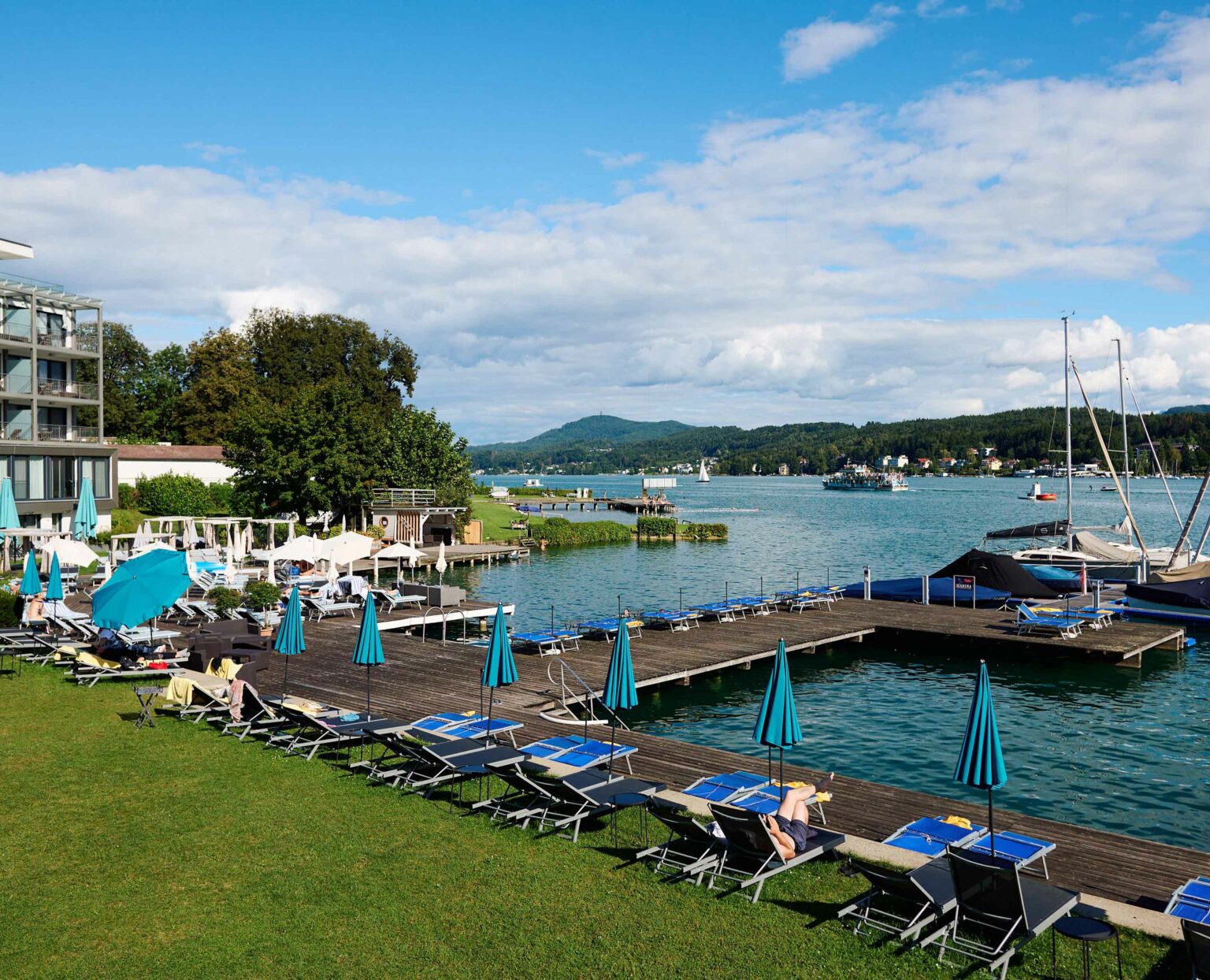 Liegewiese mit Sonnenschirmen und Badesteg am Wörthersee, moderne Hotelanlage im Hintergrund.