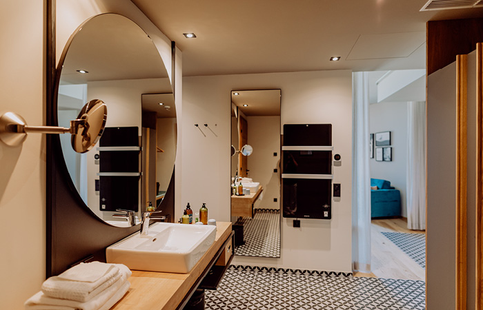 Badezimmer mit rundem Spiegel, Waschbecken auf Holzablage, schwarz-weiß gemustertem Boden und Blick in angrenzenden Raum mit Sofa