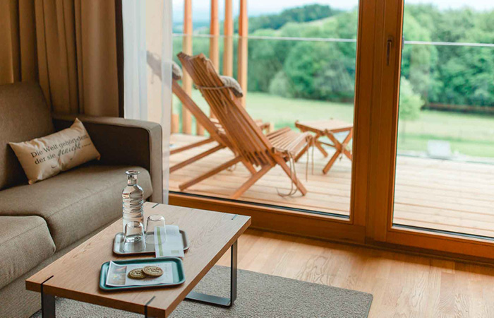 Wohnzimmer mit Sofa und Kissen mit der Aufschrift 'Der Welt geschenkt, der sie genießt', Holztisch mit Wasserflasche, Gläsern und Keksen, Blick auf Terrasse mit Holzliegen und grünem Garten