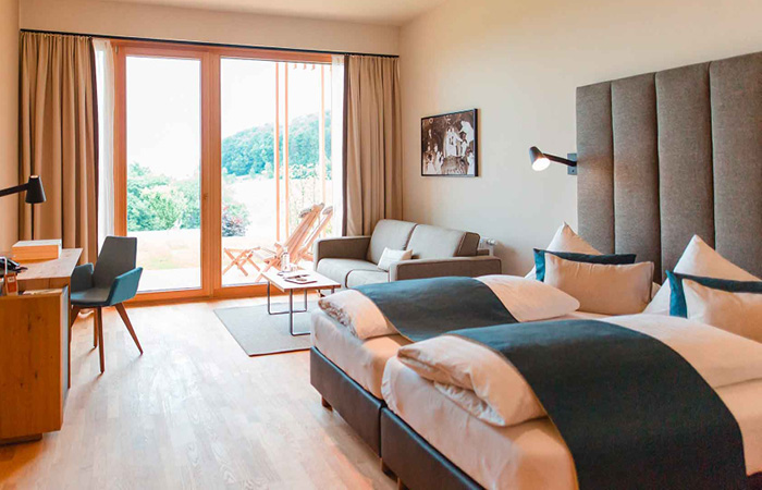Hotelzimmer mit zwei Betten, grauen Kopfteil, Sofa, Schreibtisch und großer Glasfront mit Blick auf Terrasse und grüne Landschaft