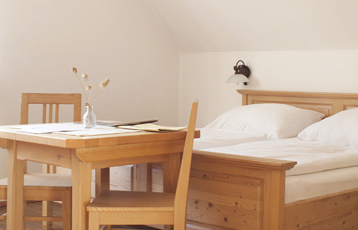Holztisch mit zwei Stühlen, Vase mit Trockenblumen, im Hintergrund ein Bett mit weißen Kissen und Decken, Wandlampe darüber