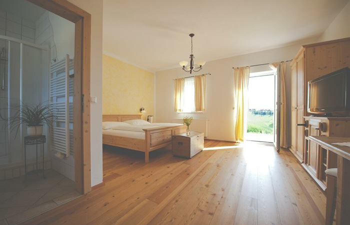 Hell eingerichtetes Schlafzimmer mit Holzboden, Doppelbett, Fenster mit Vorhängen und offenem Balkon mit Blick ins Grüne