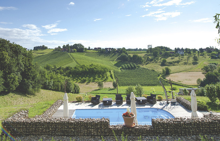 Blick auf einen rechteckigen Pool mit Liegestühlen und Sonnenschirmen, umgeben von einer Steinmauer, im Hintergrund grüne Hügel und ein bewölkter Himmel.