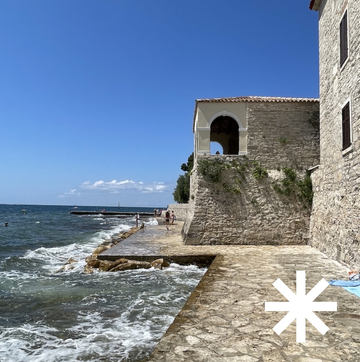 Steinmauer mit Bogenfenster am Meer, Wellen schlagen gegen gepflasterten Uferweg unter blauem Himmel, rechts unten ein Stern-Icon für die Kategorie Business-Reisen