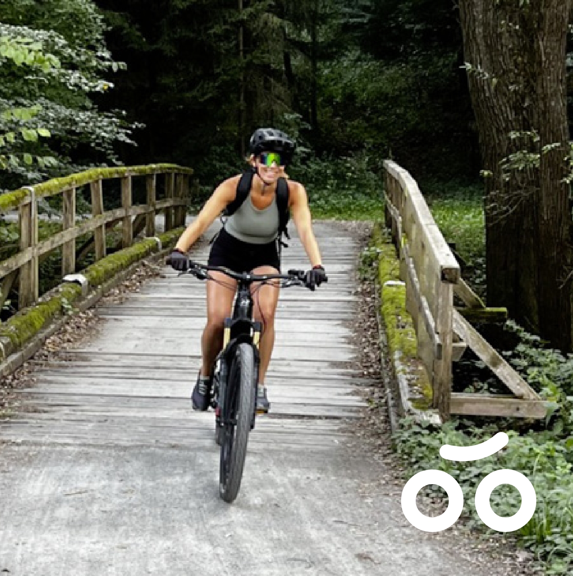Frau mit Fahrradhelm und Rucksack fährt auf einem Holzsteg durch einen Wald. Rechts unten ein Bike-Icon für die Kategorie Bike-Reisen