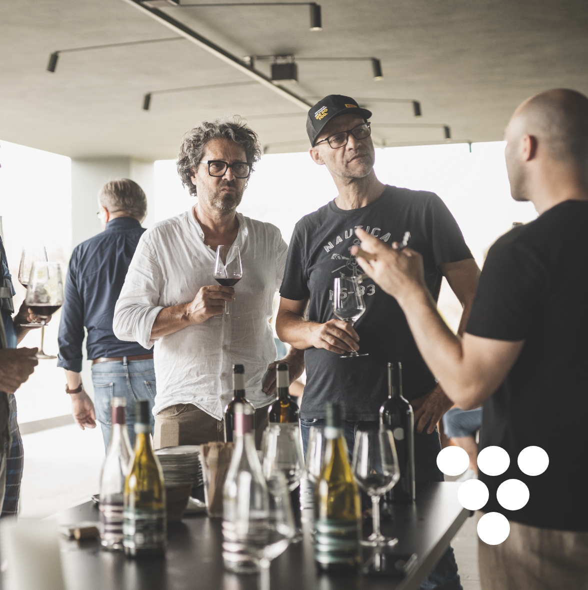 Mehrere Personen stehen um einen Tisch mit Weinflaschen und Weingläsern, einige halten Weingläser in der Hand. Rechts unten ein Trauben-Icon für die Kategorie Wein-Reisen