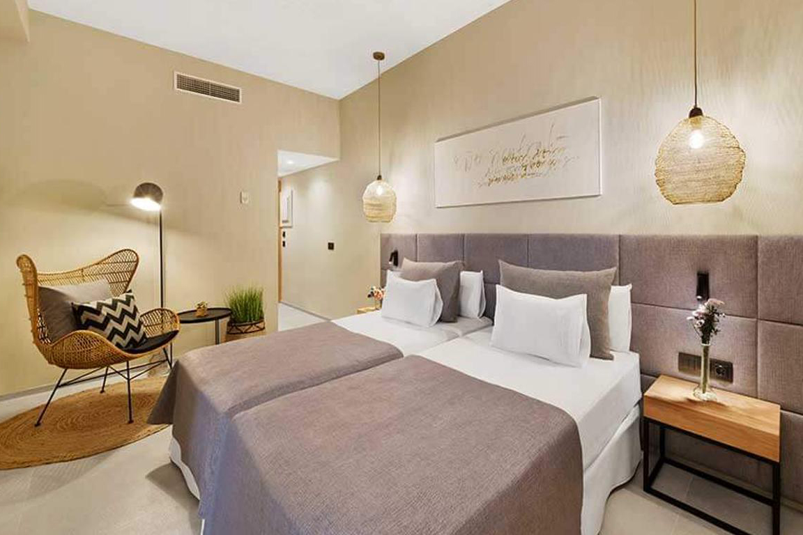 Modern eingerichtetes Hotelzimmer mit zwei Einzelbetten, sanften Naturtönen, geflochtenem Sessel und hängenden Lampen.