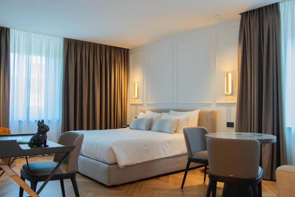 Helles Hotelzimmer im Ambassador Palace Hotel mit Doppelbett, Sitzbereich und bodentiefen Fenstern.