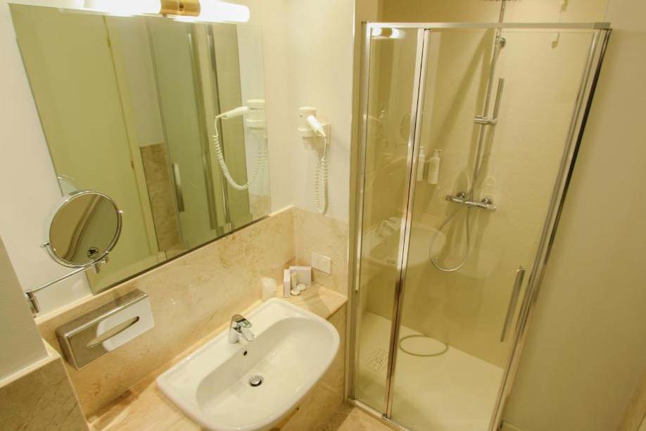 Badezimmer im Ambassador Palace Hotel mit Dusche, Waschbecken, Spiegel und hellen Fliesen.