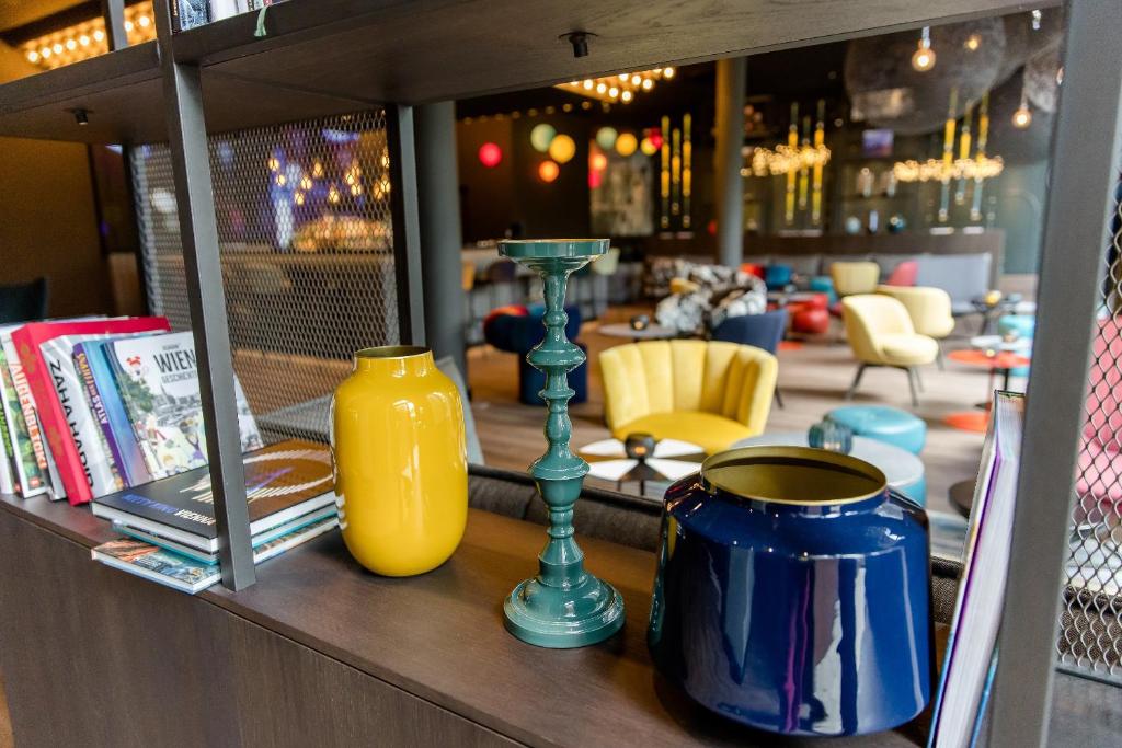 Detailansicht eines Designregals im Motel One Wien Prater mit farbigen Vasen, Kerzenständern und Blick auf die Lounge.