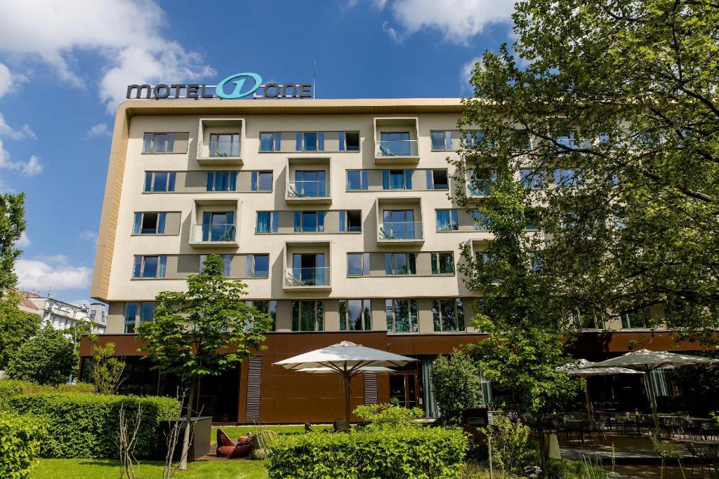 Außenansicht des Motel One Wien Prater mit Gartenbereich, Sonnenschirmen und Hotelgebäude.