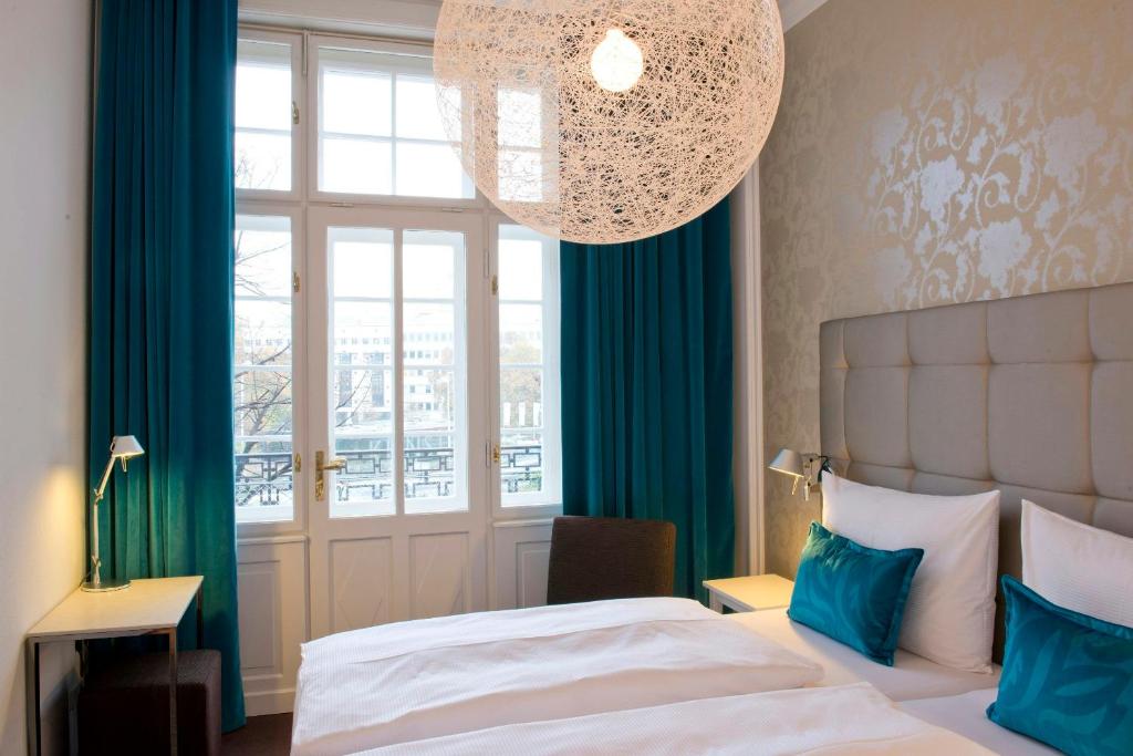 Helles Hotelzimmer im Motel One Wien Staatsoper mit Doppelbett, Balkonfenster, Design-Leuchte und Blick auf die Wiener Innenstadt.